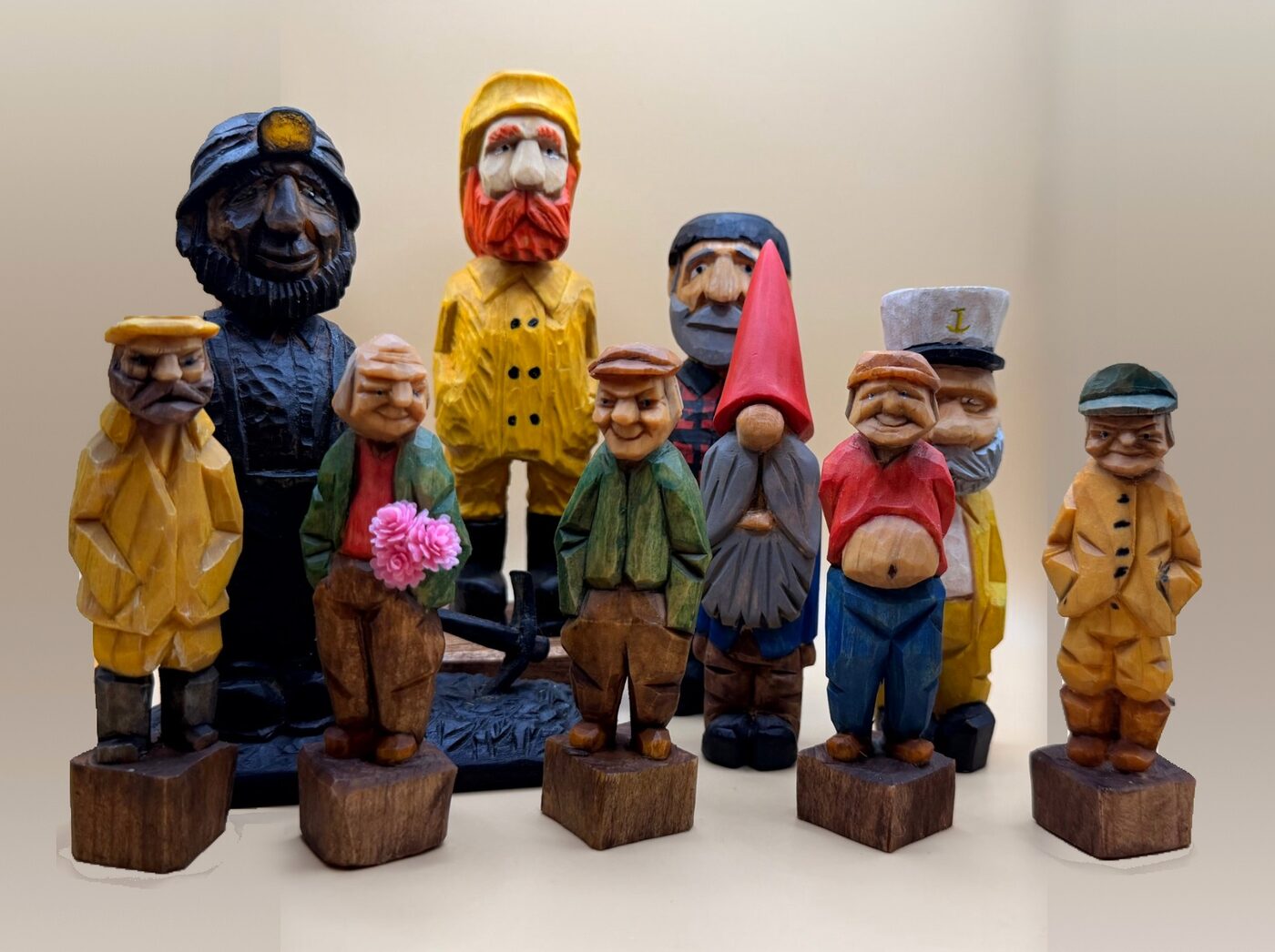 Ten hand-carved, painted wood figures standing together: a coal miner with a headlamp and pick, fishermen in yellow slickers, a man holding pink flowers, a lumberjack in red plaid, a gnome with a tall red hat, a sea captain, and a smiling man with his belly showing.